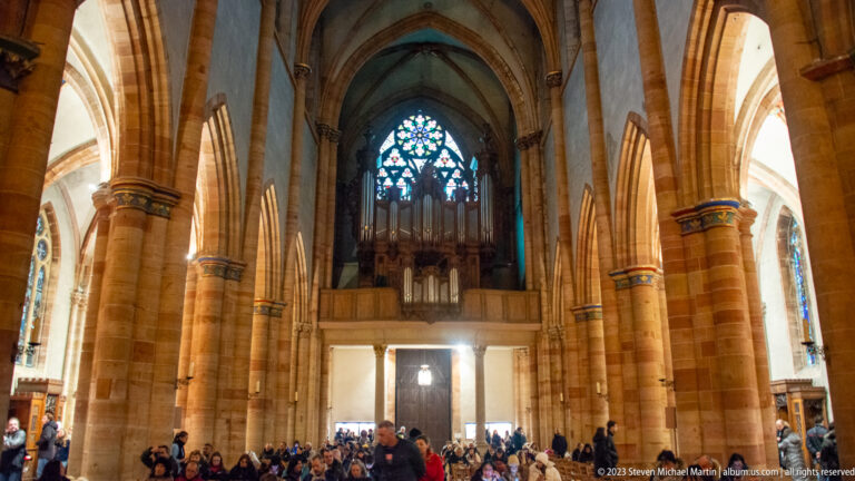 Tall Gothic nave with clustered stone piers, pointed arches, organ loft, stained glass, and visitors below.