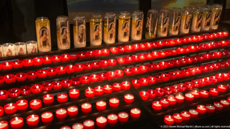 Rows of glowing red votive candles beneath devotional image candles in a dim church interior.