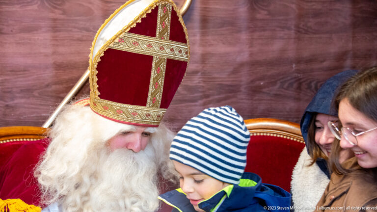 White-bearded Saint Nicholas in a red-and-gold mitre greeting a child while visitors gather beside him.