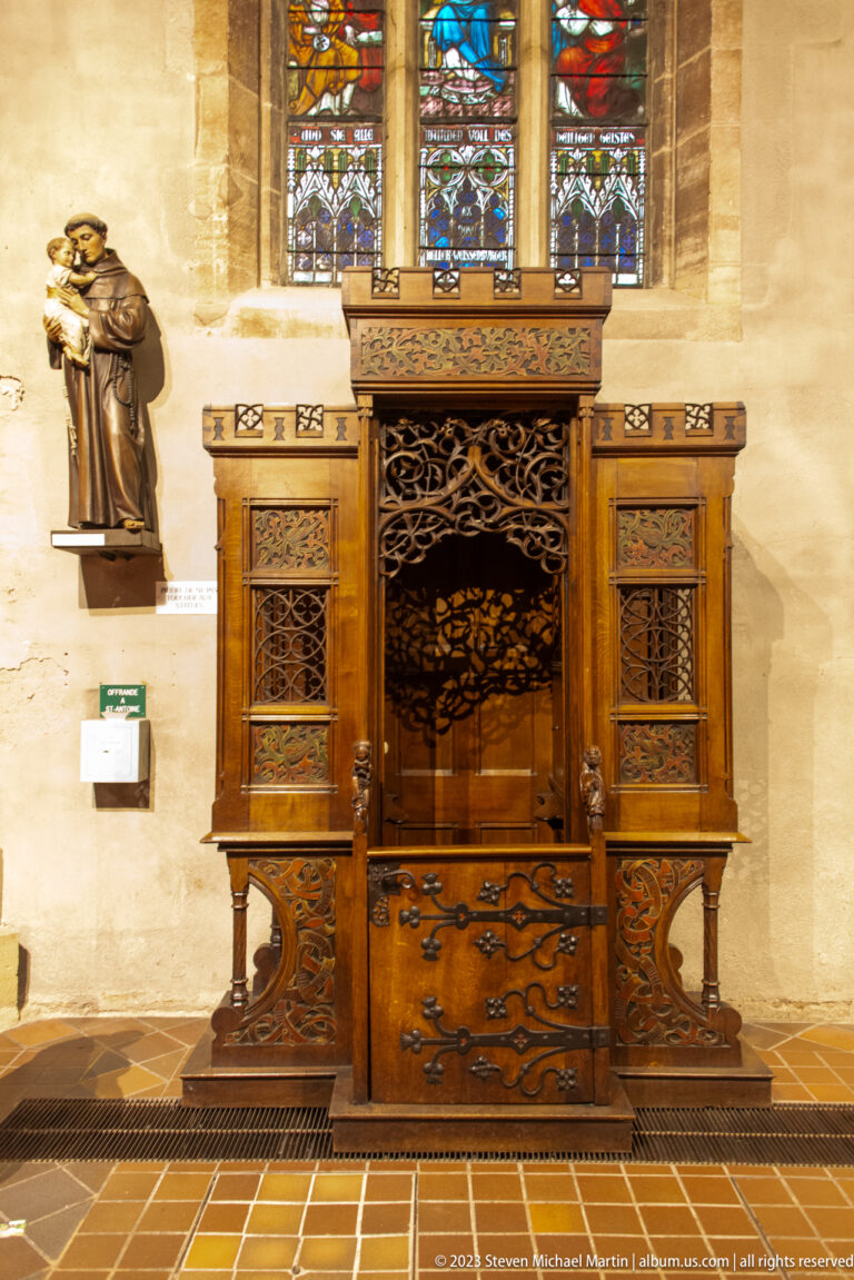 Ornately carved dark wooden church furnishing with pierced panels beneath stained-glass windows and beside a religious statue.