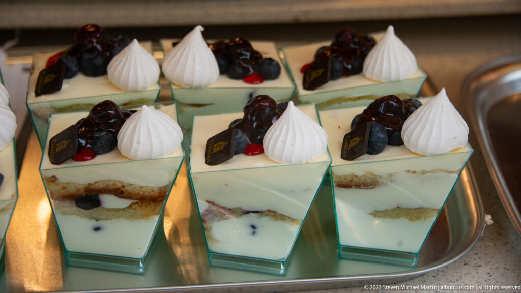 Six layered square pastries on a metal tray, topped with berries, meringue kisses, and small chocolate plaques.