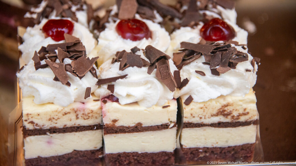 Square layered cakes topped with whipped cream, red cherries, and chocolate shavings in a close bakery display.