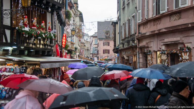 Crowded Colmar street in rain, filled with umbrellas beneath decorated historic façades and Christmas lights.