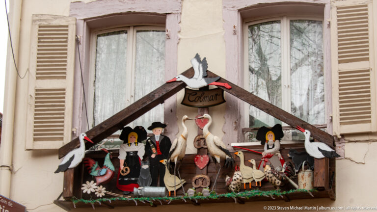 Decorative window box labeled Colmar with storks, hearts, flowers, and small figures in traditional-style dress.