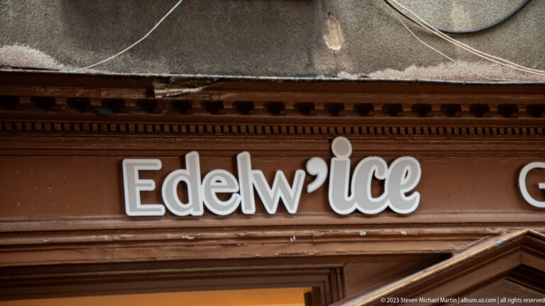 Close view of raised white Edelw'ice lettering on a dark brown storefront fascia beneath a traditional cornice.