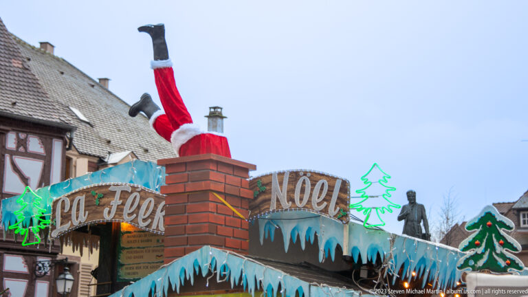 Santa figure upside down in a mock chimney above a Christmas stall decorated with icicles and “NOEL” lettering.