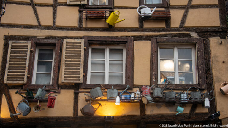 Half-timbered façade with shuttered windows and rows of watering cans, pots, baskets, and other objects mounted outside.