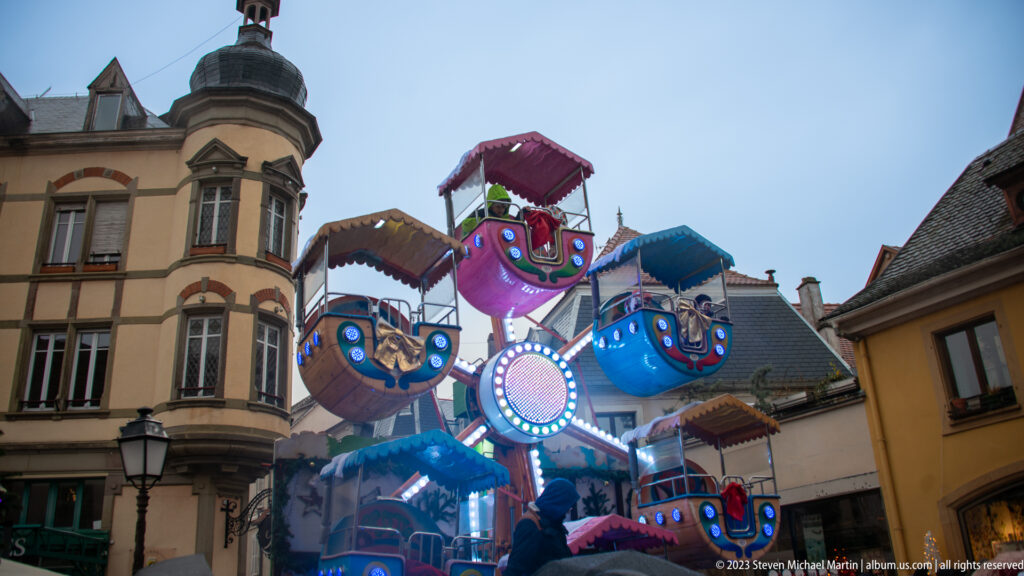 Colorful illuminated children's fairground ride with small gondolas and canopies between historic Colmar buildings.
