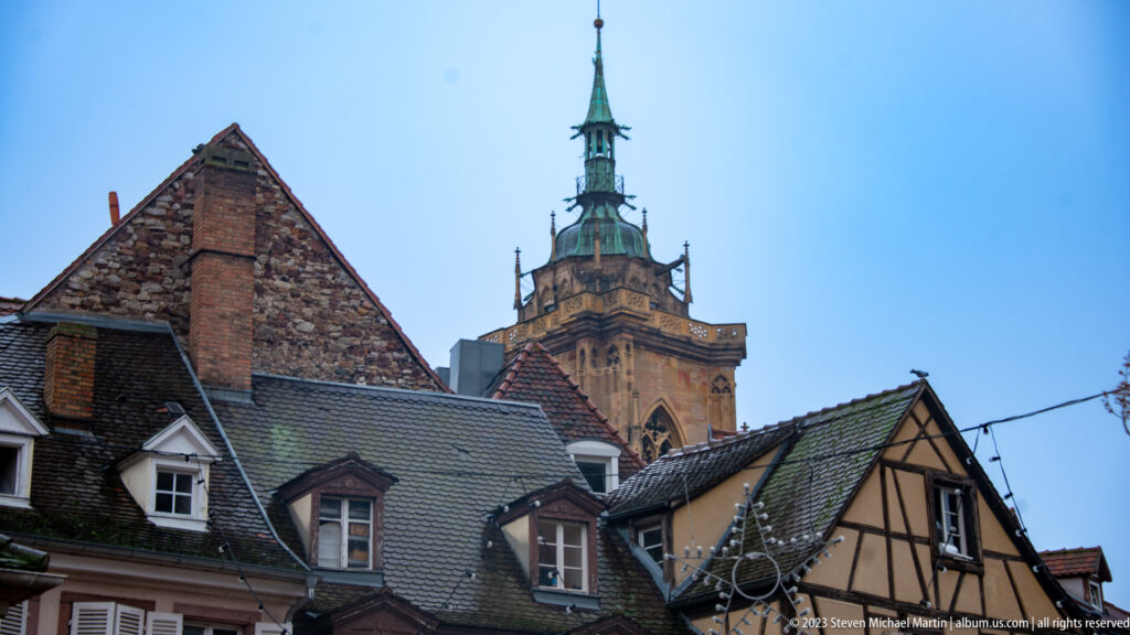 Saint Martin's Church tower and green spire rising above tiled roofs, dormers, chimneys, and half-timbered buildings.