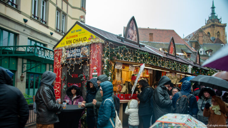 Rainy Christmas-market crowd beside a decorated stall advertising vin chaud and crêpes in Colmar.