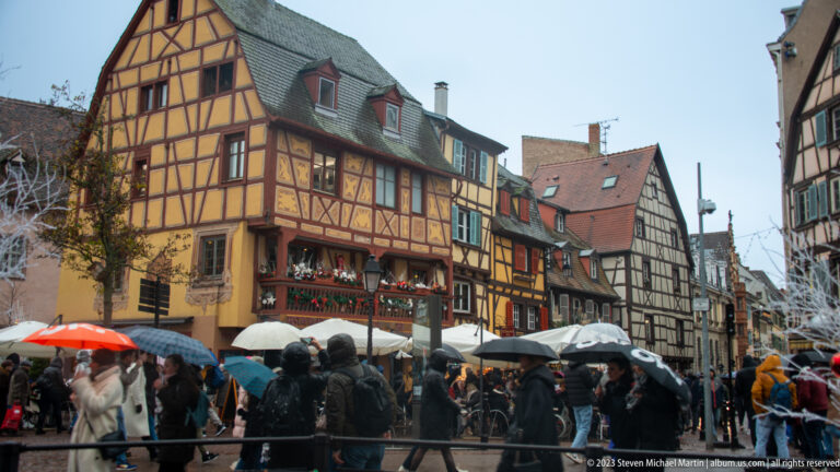 Crowded rainy street lined with colorful half-timbered houses, umbrellas, garlands, and Christmas decorations in Colmar.