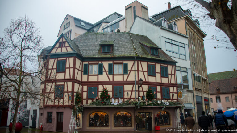 Small half-timbered corner house with shutters, steep roof, Christmas greenery, and lit shop windows below.