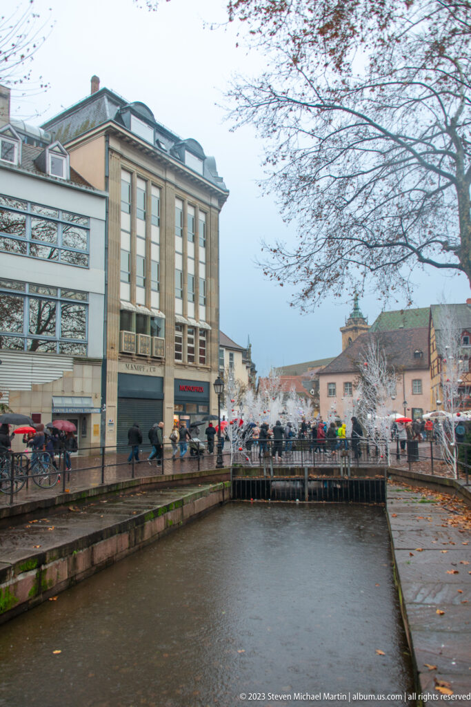 Canal in Colmar leading toward a low footbridge, with buildings, bare trees, and rainy pedestrians along the banks.