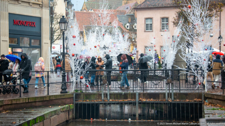 White decorative trees with red ornaments beside a crowded pedestrian bridge over a Colmar canal.