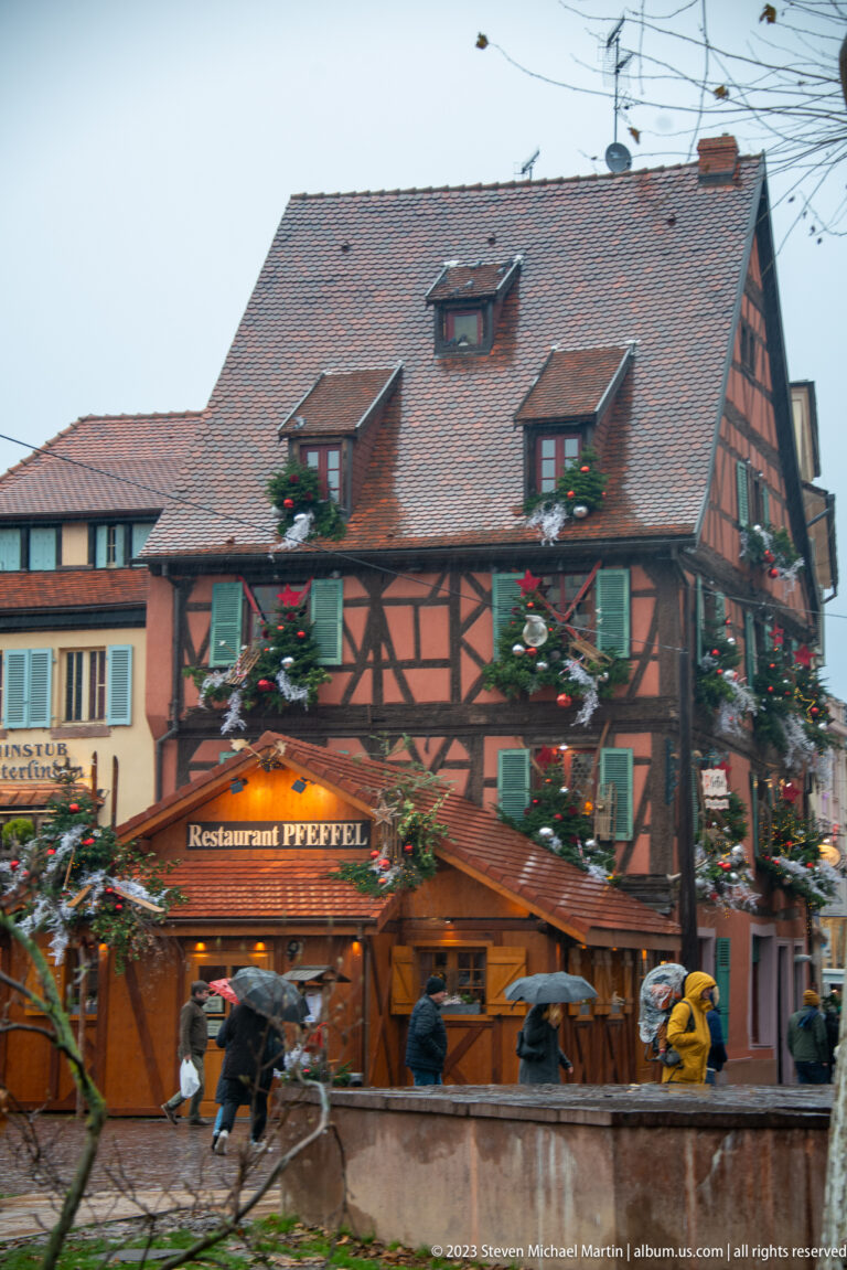 Half-timbered Restaurant Pfeffel decorated with evergreen garlands beside rainy pedestrians in Colmar.