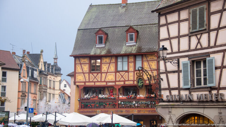 Decorative ochre-and-red half-timbered building above white Christmas-market umbrellas in a narrow Colmar street.