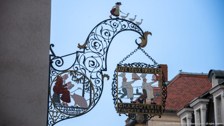 Ornate hanging sign reading Aux Deux Frères and Charcuterie Fincker Frères with painted figures and metal scrollwork.