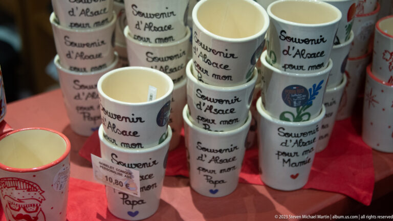 Stacks of small cream ceramic cups lettered Souvenir d'Alsace with family dedications such as pour mon Papa.