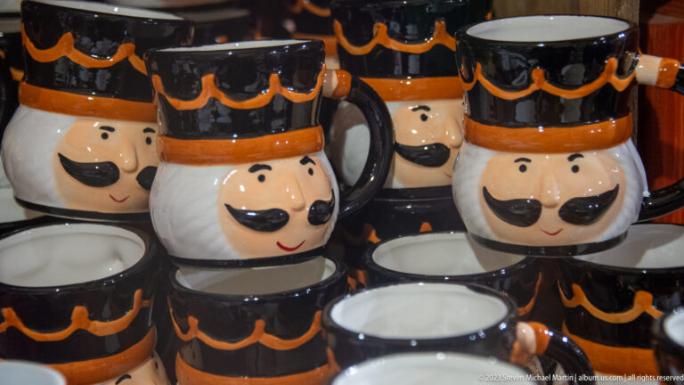 Rows of ceramic souvenir mugs shaped as smiling moustached faces with black-and-orange hats.
