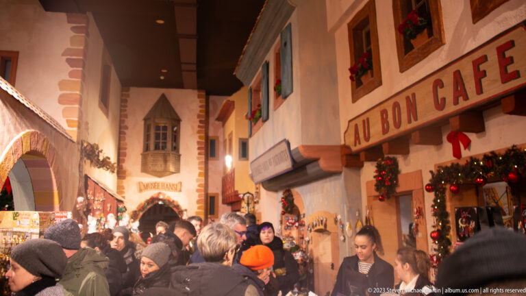 Crowded evening street beside Au Bon Café, with Christmas garlands, wreaths, lights, and market visitors.