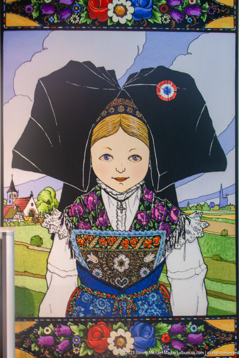 Decorative illustration of a woman in Alsatian-style dress with a large black headdress, floral patterns, and rural background.
