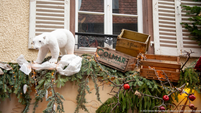 White polar-bear figure beside a rustic “COLMAR” sign, evergreen branches, artificial snow, and red ornaments.