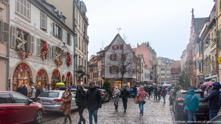 Wet cobbled Colmar street with pedestrians, decorated historic façades, parked cars, and a gabled house ahead.