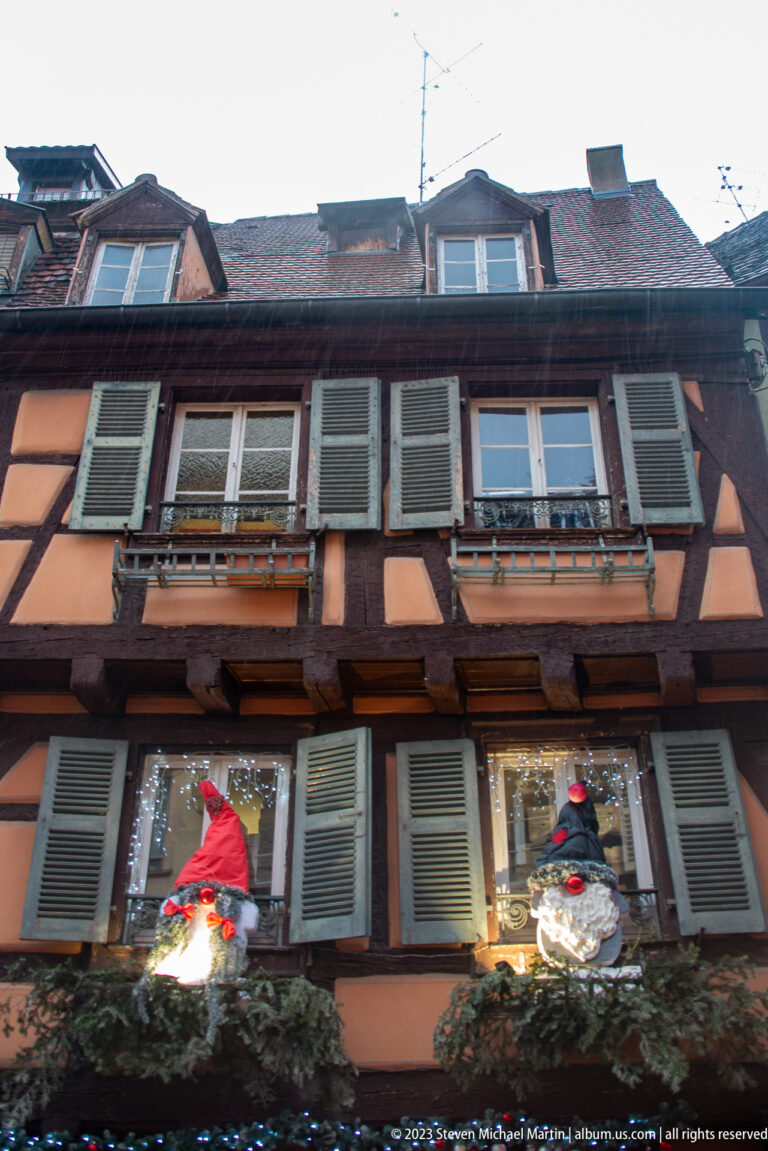 Salmon-colored half-timbered house with green shutters, flower boxes, and illuminated Christmas figures beneath a tiled roof.