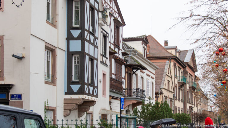 Narrow Colmar street with half-timbered corner house, shutters, balconies, tiled roofs, and bare winter trees.