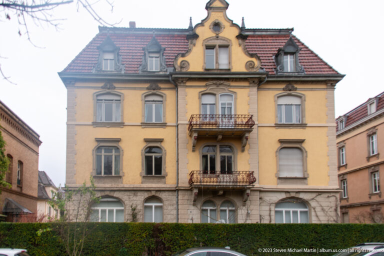 Yellow historic villa with ornate central gable, dormer windows, stone trim, balconies, and hedge in front.