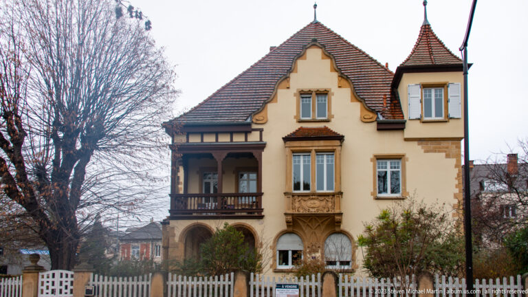 Cream historic villa with timber loggia, arched opening, projecting bay, decorative stonework, and steep tiled roofs.