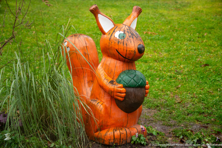 Painted wooden squirrel sculpture holding a large acorn beside ornamental grass on a green lawn.