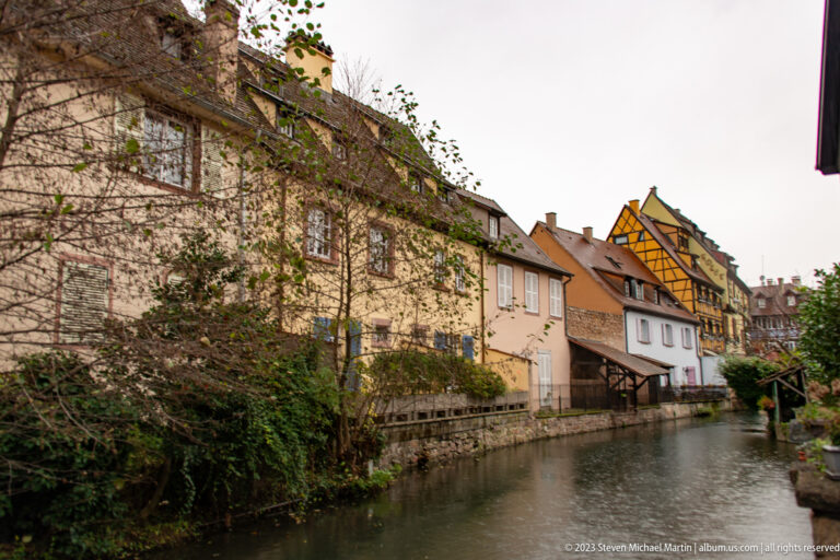 Quiet Colmar canal bordered by pale houses, vegetation, and a yellow-brown half-timbered building in the distance.