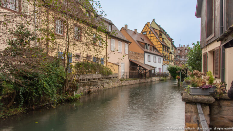 Centered canal view toward traditional houses, with stone edges, planters, and a yellow half-timbered façade ahead.
