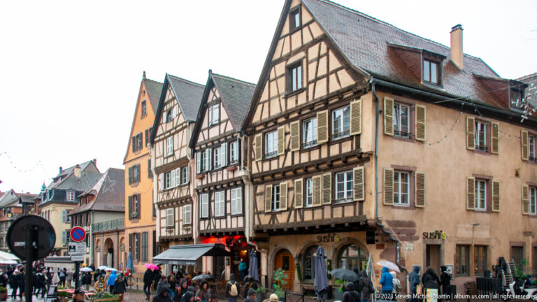 Tall half-timbered houses with steep gables and shops lining a crowded pedestrian street in Colmar.
