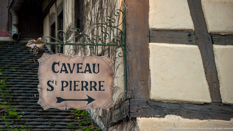 Metal Caveau St Pierre sign with arrow suspended from ornate ironwork on a half-timbered wall.