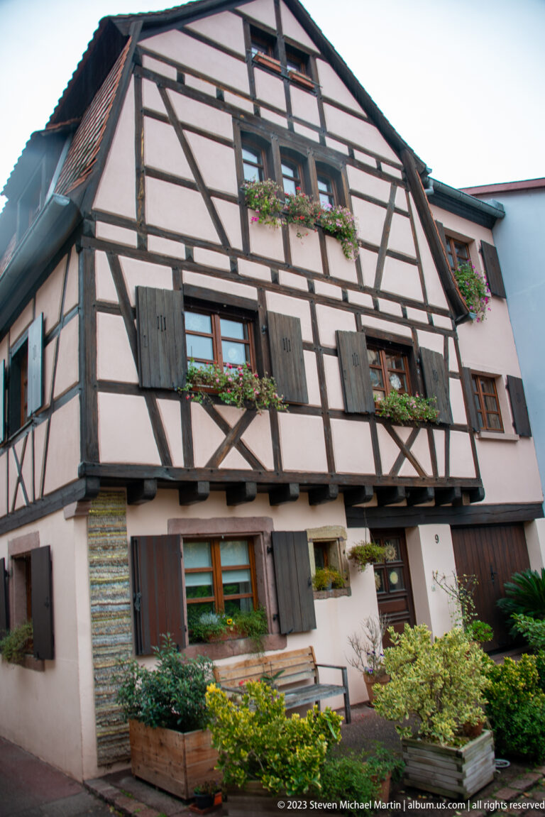 Large half-timbered house with dark beams, shuttered windows, flower boxes, and projecting upper story.