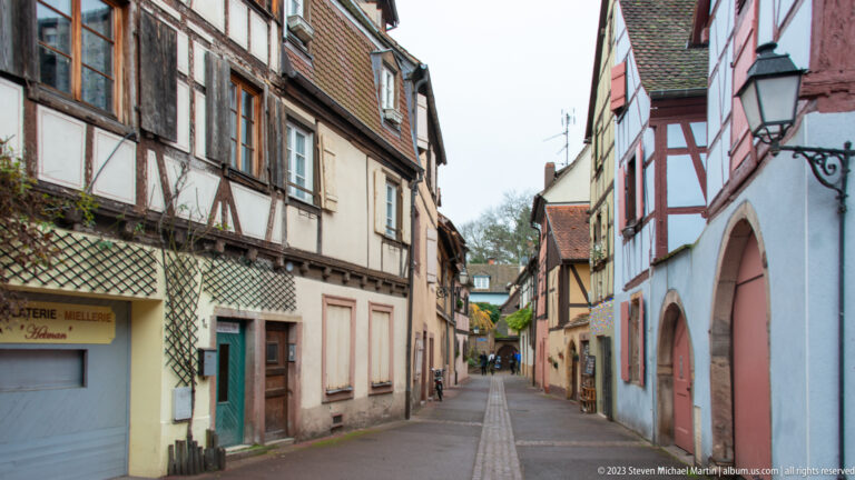 Narrow Colmar lane with timber-framed and plastered houses, shutters, arched doorways, and projecting signs.