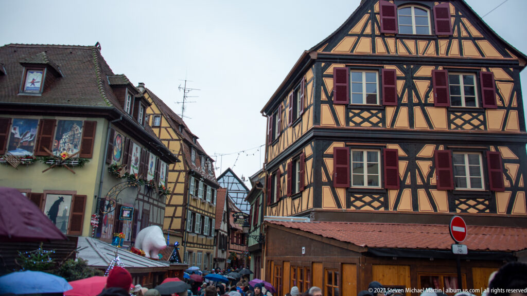 Crowded Colmar Christmas-market street beneath tall decorated half-timbered houses with painted shutters.