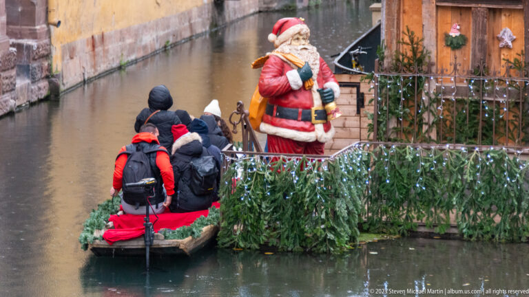 Visitors in a small canal boat pass a large Santa figure and evergreen garlands beside the water.