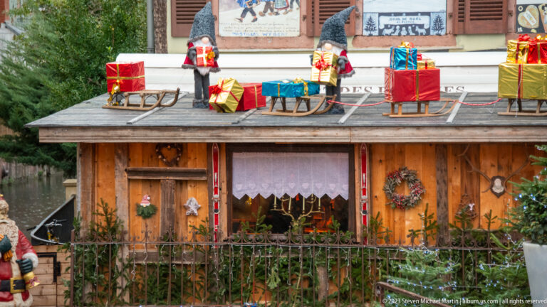 Wooden Christmas-market hut topped with gnomes, miniature sleds, wrapped gifts, greenery, and a wreath.