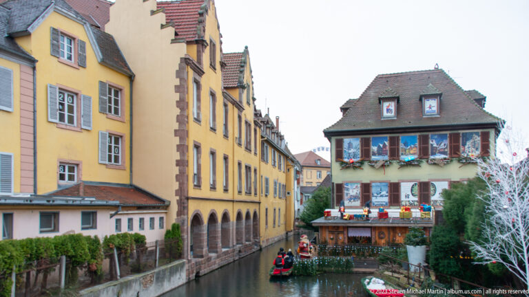 Canal between yellow historic buildings, with Christmas decorations, market structures, and a small boat in the distance.
