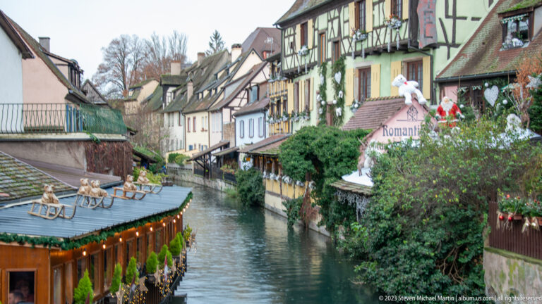 Colmar canal lined with half-timbered houses, pitched roofs, greenery, and decorated Christmas-market structures.