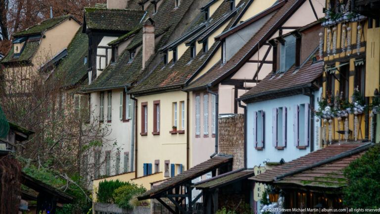 Closely packed canal-side houses with steep tiled roofs, dormers, chimneys, pastel walls, and timber framing.