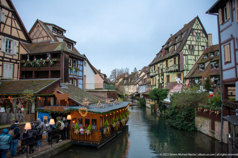 Wide Colmar canal view with half-timbered houses, decorated wooden market stall, and visitors along the bank.