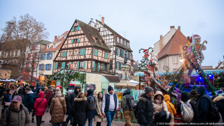 Large Christmas-market crowd below half-timbered houses, illuminated decorations, and a tall whimsical holiday figure.