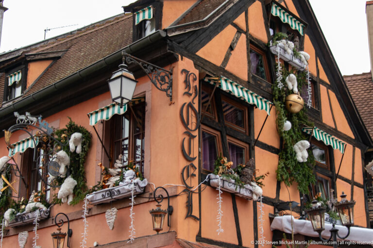 Orange half-timbered corner building with evergreen garlands, white animal figures, baubles, striped awnings, and icicle lights.