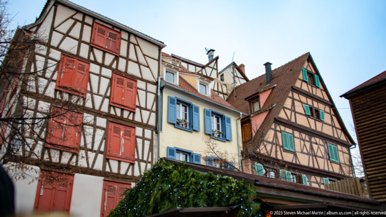 Three colorful half-timbered houses with red, blue, and green shutters beneath steep tiled roofs.