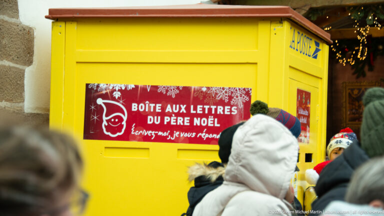 Bright yellow Santa mailbox with French sign reading Boîte aux lettres du Père Noël, surrounded by winter-clad visitors.
