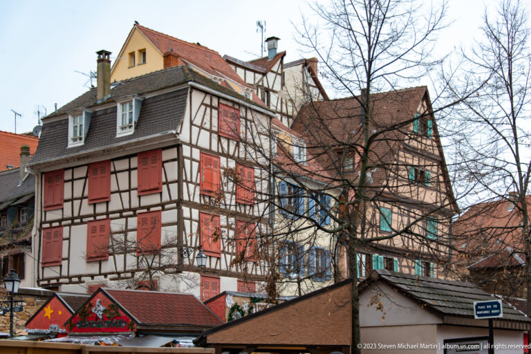 Half-timbered houses with red shutters and steep roofs rising above low Christmas-market structures and bare branches.